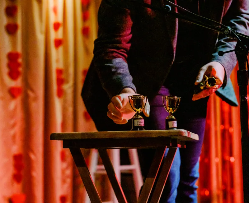 Two small trophies on a stool in front of a red curtain, a host reaching for one