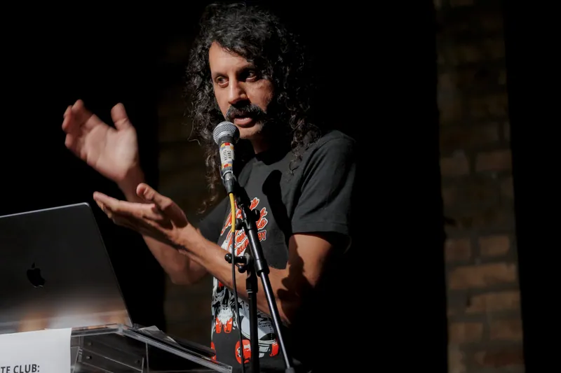 A combatant gesturing with both hands, laptop open on the lectern