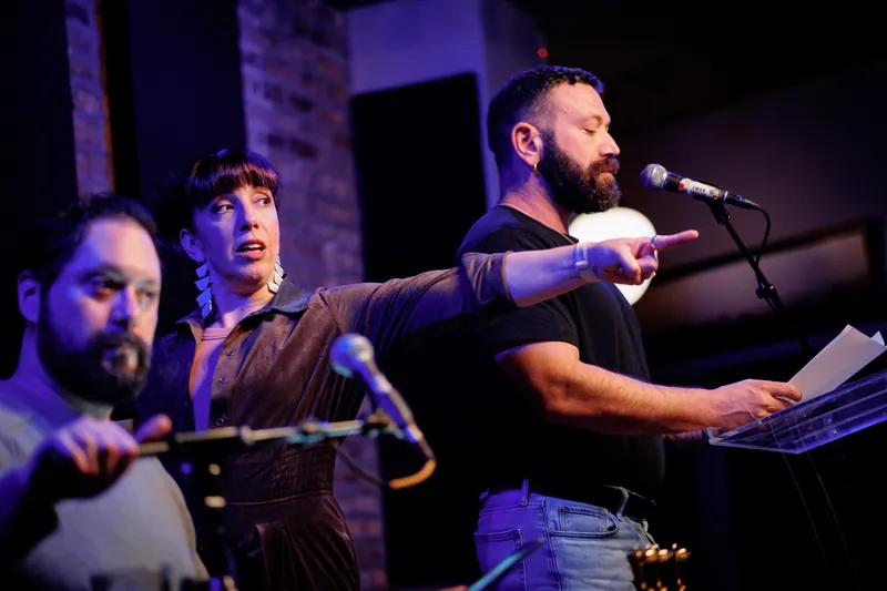 Three performers at the microphones, one pointing out at the room