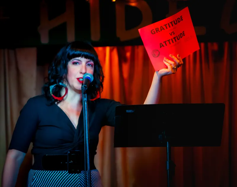 A combatant on stage with a GRATITUDE versus ATTITUDE card held up beside her