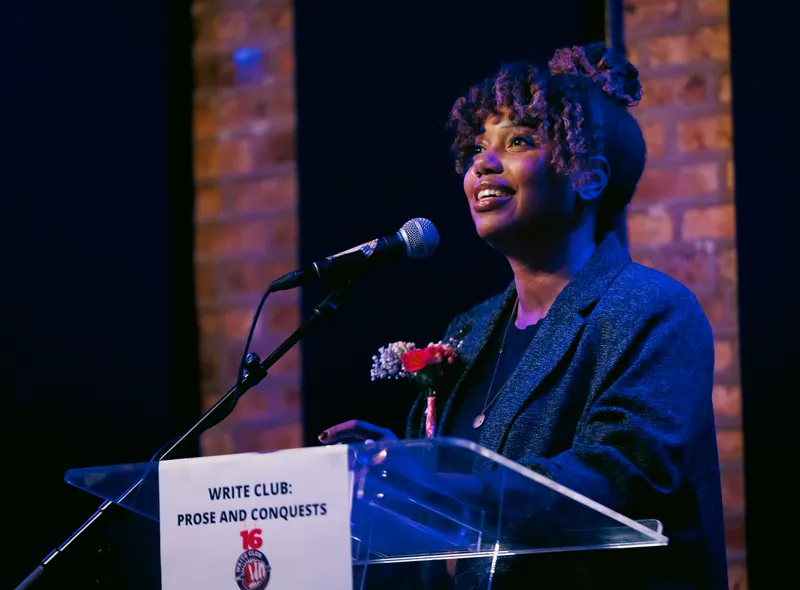 A combatant smiling at the lectern, the Prose and Conquests sign below her