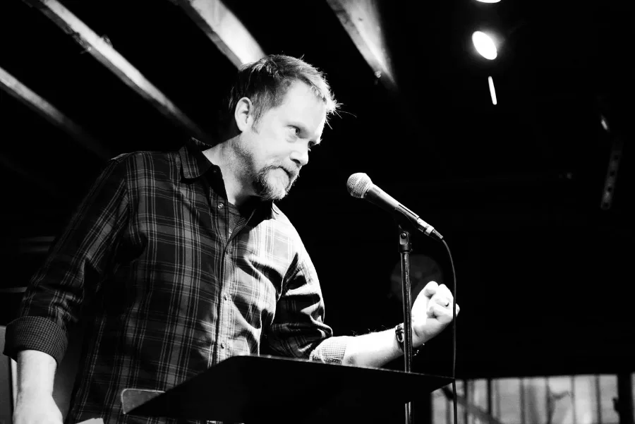 Ian Belknap at a lectern with a microphone, mid-reading